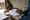 A person reviews a printed chart beside blister packs while a woman sits at a table indoors.