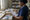 A woman in scrubs checks a handwritten list beside boxes of packaged supplies in a storeroom.
