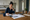 Woman writes on paperwork at a kitchen table beside a laptop, calculator, and coffee mug.