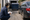 Worker crouches with a clipboard beside a dumpster, parked sedan, and garbage truck in an alley.
