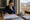 A woman studies house plans and writes at a table beside a model home and property photos.