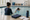 Swim instructor watches a child practicing in an indoor pool beside a clipboard, towel, and water bottle.