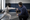 A fencing instructor crouches beside a masked fencer, pointing toward the extended sword in an indoor salle.