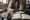 Person holding a small test device across a table while another person sits beside an open notebook.